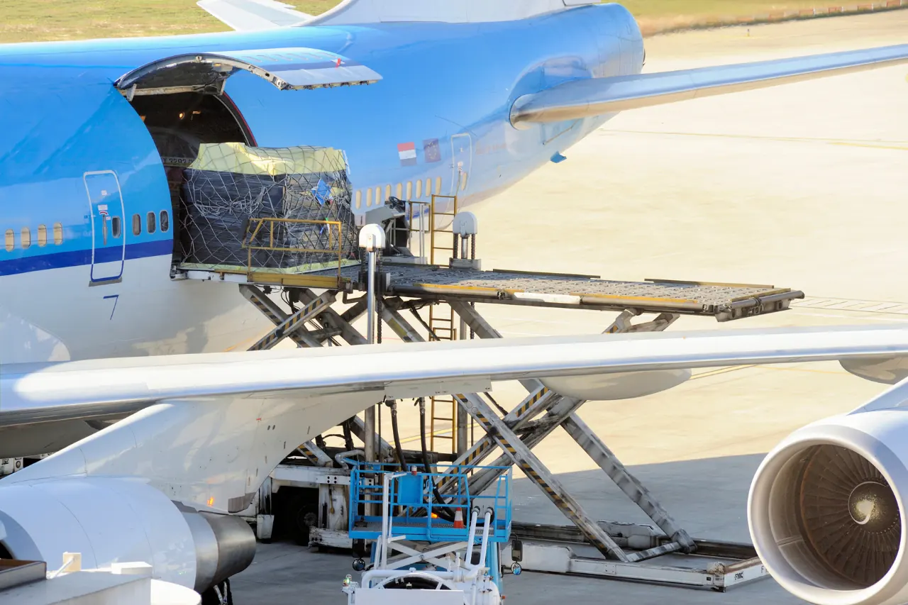 Cargo is being loaded into the side of a large blue and white airplane using a scissor lift on the tarmac, epitomizing XRT Logistics' expertise. The cargo, secured with netting on a pallet, aligns perfectly with modern freight and logistics services. The airplane's engine and wing are visible in the foreground.