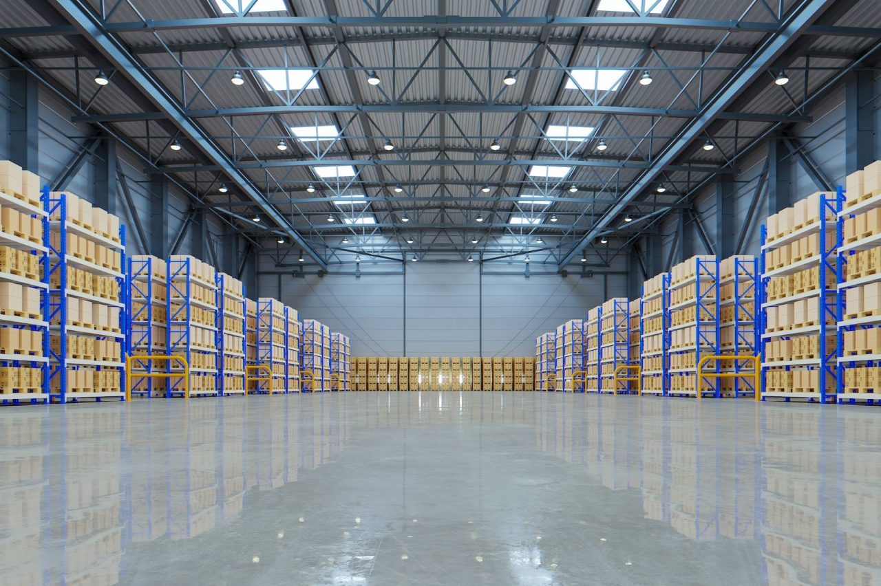 Interior of a spacious, modern warehouse with high ceilings. Shelves line the walls, filled with neatly stacked cardboard boxes. The floor is polished concrete, reflecting the overhead lighting.