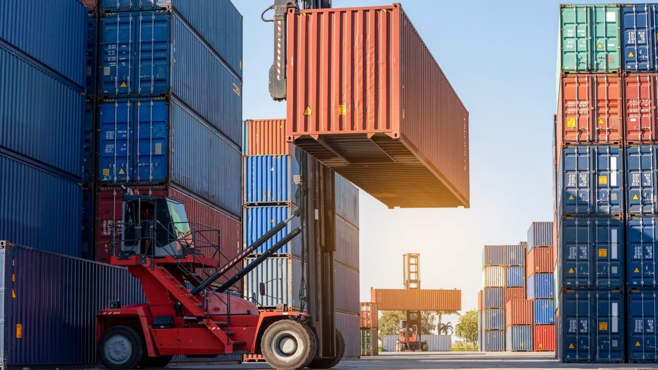 A red forklift is lifting a large orange shipping container amidst stacks of blue and multicolored containers in a shipping yard. The sky is clear and the sun casts long shadows on the ground.