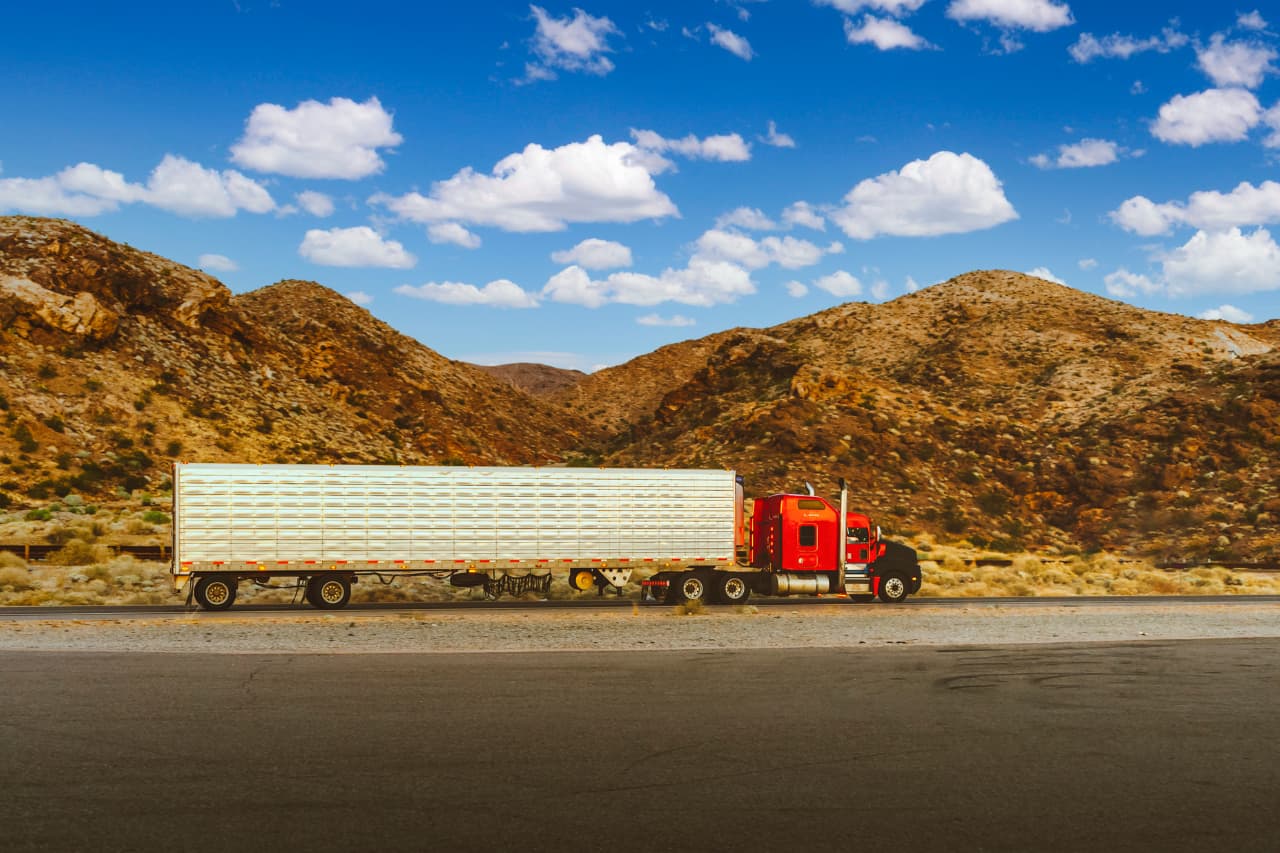 A red semi-truck with a large white trailer, bearing the hallmark of freight and logistics services, is parked on a road. Behind it, a rocky desert landscape and hills stretch into the distance under a clear blue sky with scattered clouds.