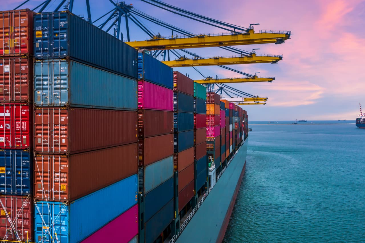 A large cargo ship loaded with colorful shipping containers, representing XRT Logistics, is docked at a port. Yellow cranes hover above as the sun sets over the calm sea, painting the sky in vibrant hues of pink and blue—a perfect scene for international transportation solutions.