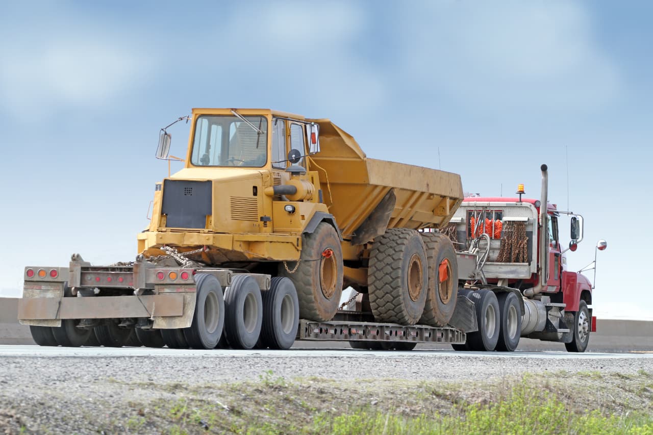 A large red truck from XRT Logistics is expertly hauling a yellow construction vehicle on a flatbed trailer along the roadway. The sky is clear and blue, showcasing seamless freight and logistics services in action.