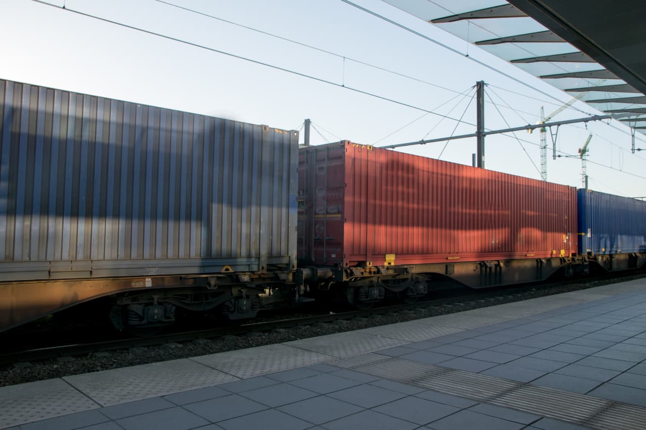 The freight train, featuring blue and red shipping containers, moves methodically along a railway track at an empty station. With tiled flooring beneath and power lines overhead against the clear sky, it's clear that XRT Logistics is at the forefront of international transportation solutions.