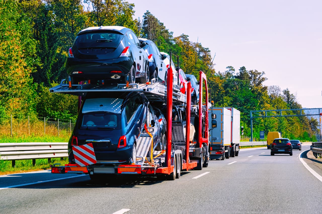 A car transporter carrying multiple cars travels on a highway, showcasing the efficiency of expert freight and logistics services. Trucks and other vehicles share the road, while trees line the way under a clear sky.