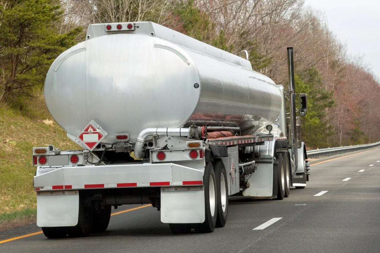 A silver tanker truck marked Oversized Load Transport drives on a highway, carrying a large cylindrical tank. The road is lined with trees sporting sparse leaves, hinting at winter or early spring. An overcast sky looms above, casting a gray hue over the scene.