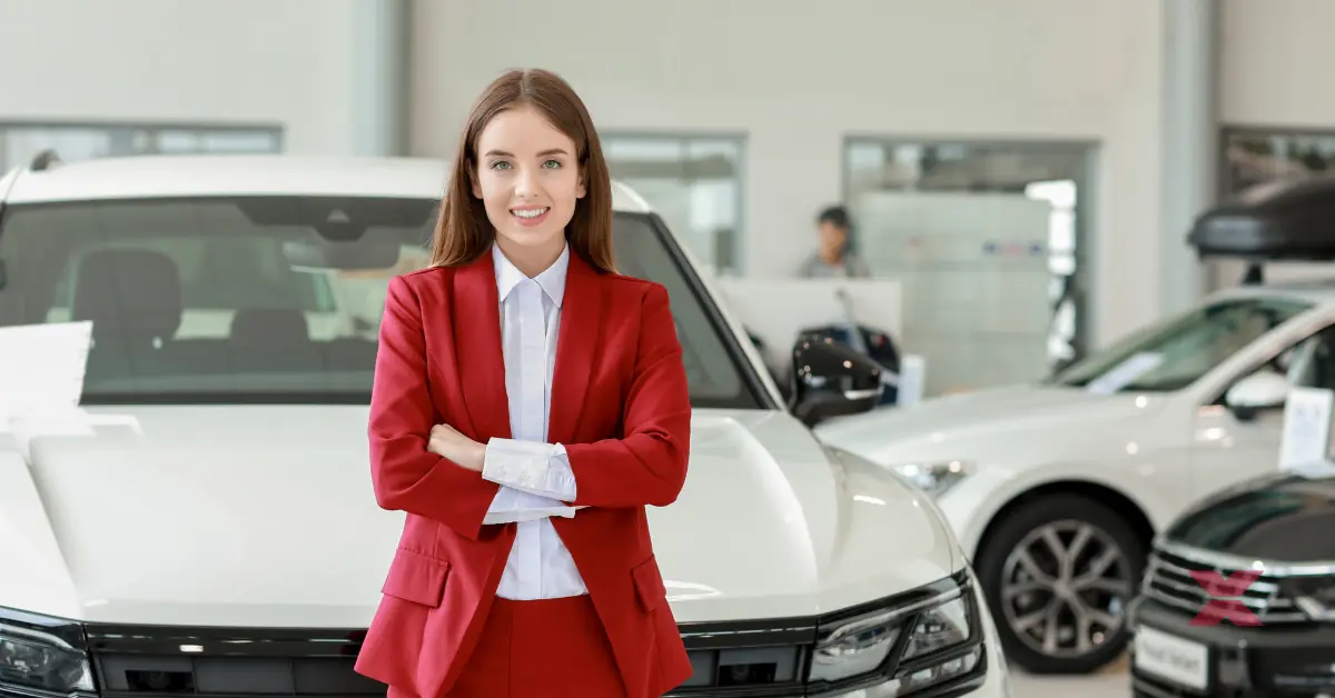 A woman in a red suit stands with arms crossed, smiling in front of a white car inside a modern car showroom, where auctioned vehicles transport services ensure seamless delivery of cars and equipment to their new owners.